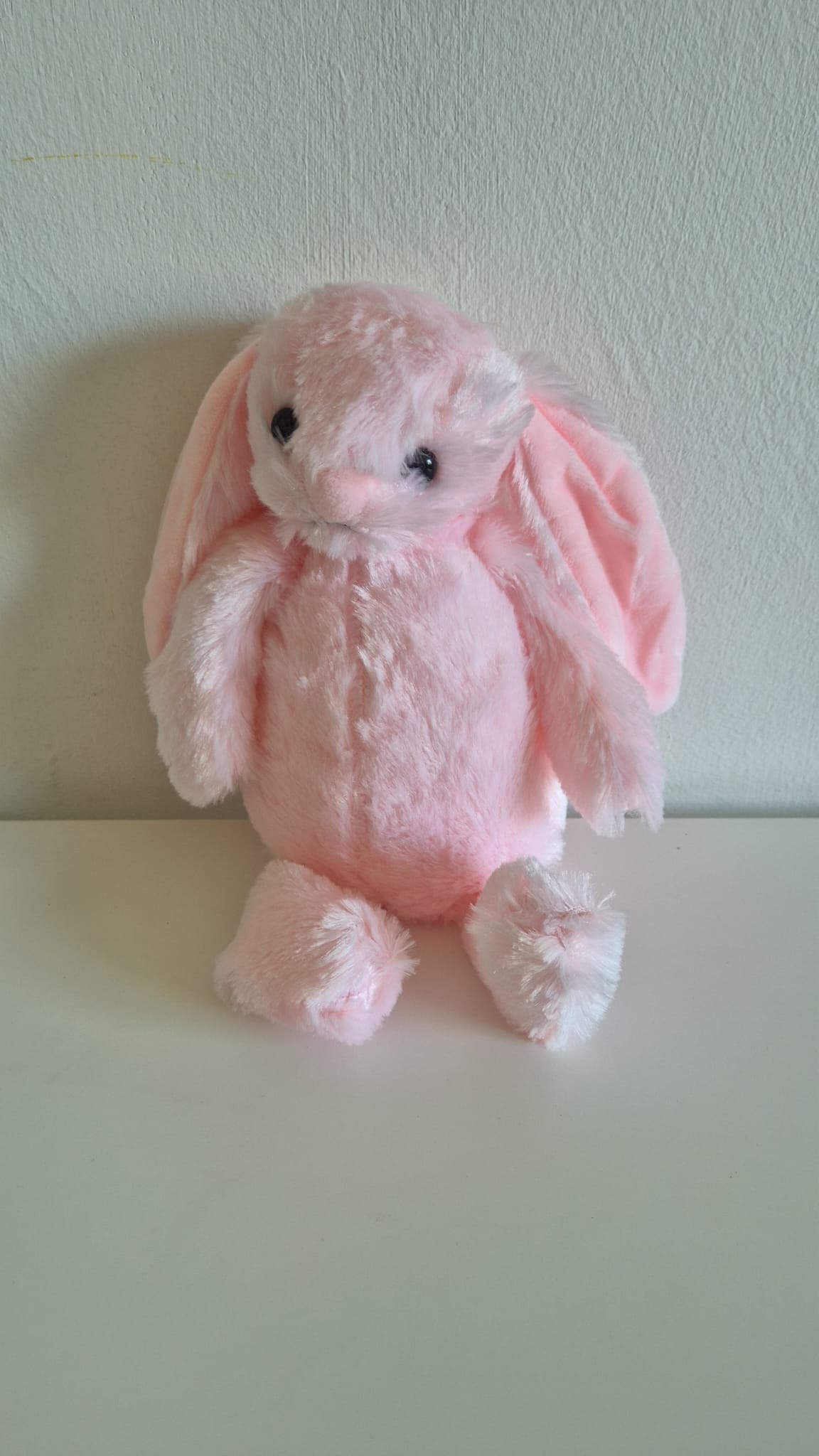 Pluche roze knuffelkonijn met lange hangoren, zachte vacht en zwarte kraaloogjes, zittend tegen een witte wand.
